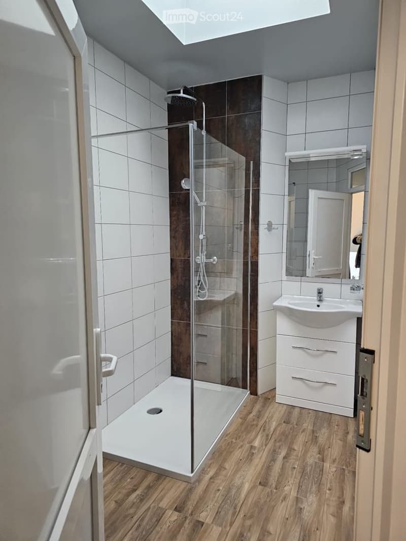 Bathroom with white tiles, wooden floor, glass shower enclosure, white sink, mirror, and silver faucet