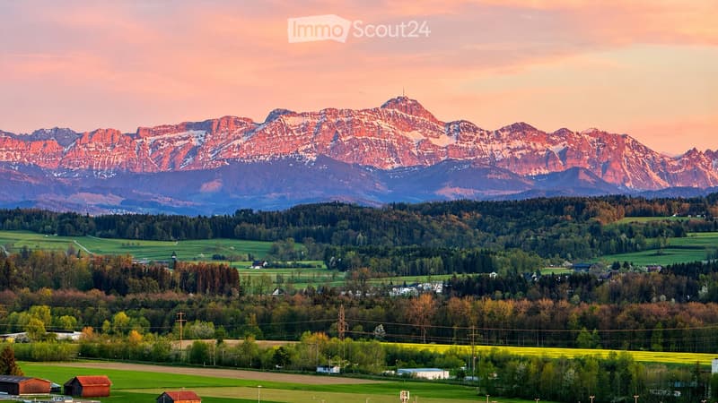A panoramic view of a lush green valley with scattered houses, surrounded by dense forests and towering mountains under a warm sunset sky.