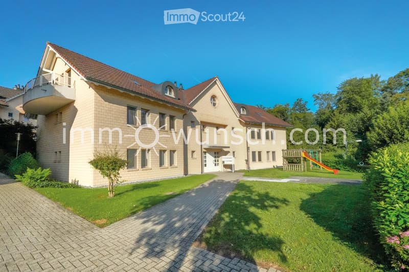 Two-story house with beige brick facade, red-tiled roof, multiple windows, and a balcony on the upper level. The house is surrounded by a well-maintained lawn, a paved walkway, and a small playground area with a slide and climbing frame.