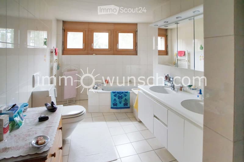 Bathroom with a toilet, bathtub, sink, mirror, and various toiletries. The room features tiled walls and floor, wooden windows, and a towel hanging on a rack.