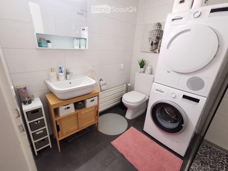 Modern bathroom with white tiles, a sink on a wooden shelf, a toilet, a washing machine, a mirror, and a small plant on the shelf.