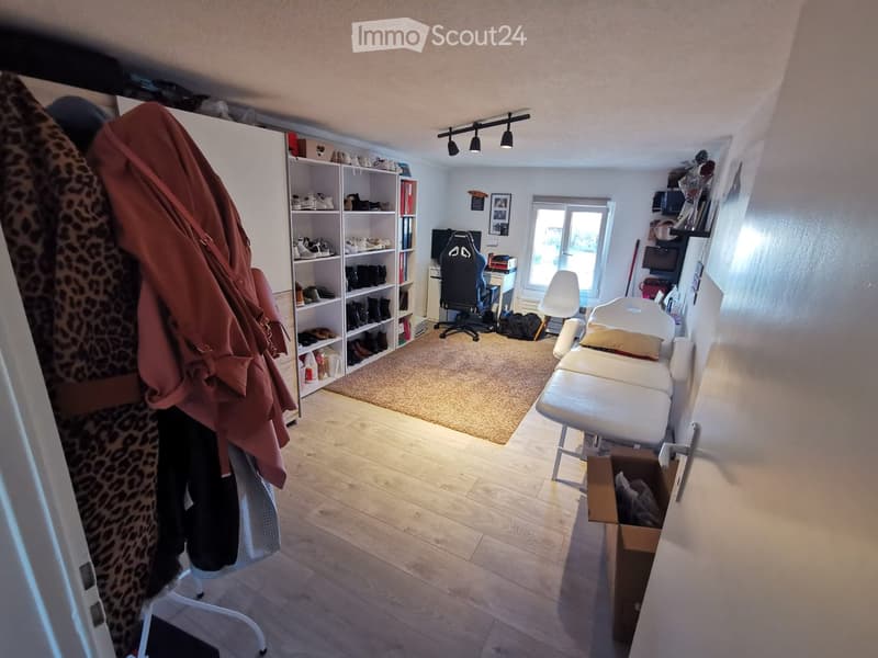 A well-lit room with wooden flooring, a large window, a shelf filled with shoes and books, a gaming chair, a small table with a screen, and a white massage table.