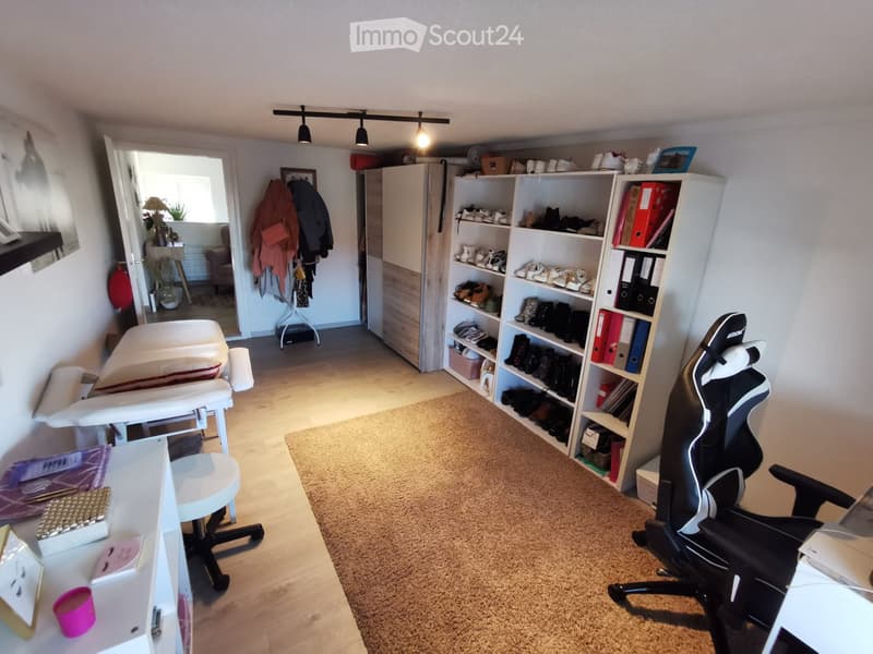 Well-lit room with a mix of furniture including a chair, table, shelves, and a mirror. The space features a carpeted area and a wooden floor.