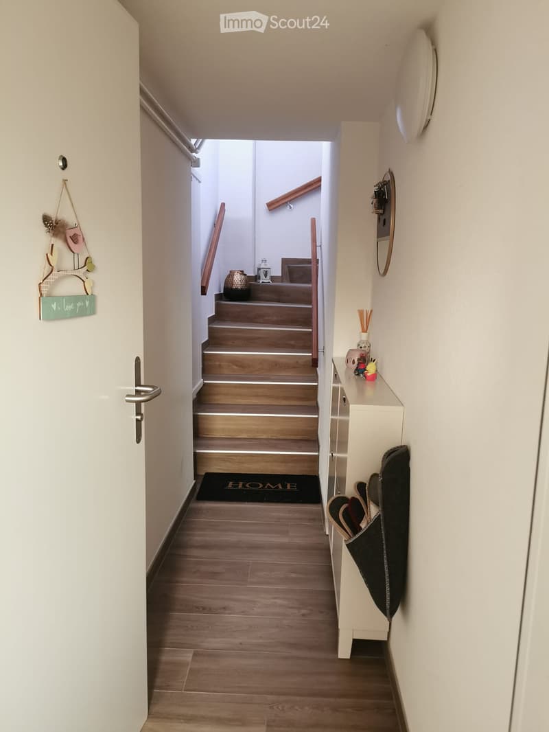 A well-lit hallway with wooden flooring, a staircase leading upwards, a small console table with decorative items, and a welcome mat at the entrance.