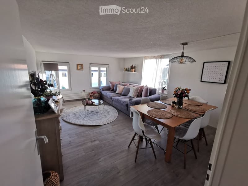 Open-plan living and dining area with a grey sectional sofa, a round coffee table, a wooden dining table with four white chairs, and decorative items including vases and a picture frame.