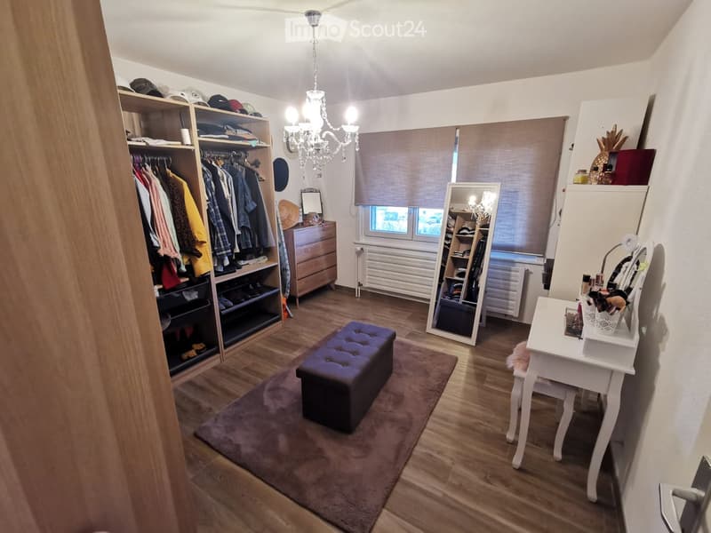 Well-lit room with wooden flooring, a large wardrobe, a vanity table with a chair, a full-length mirror, a tufted stool, and a chandelier.