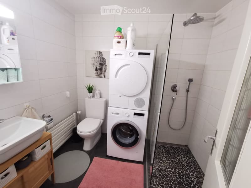 Modern bathroom with white tiles, a sink, toilet, washing machine, shower, and a small plant on the windowsill.