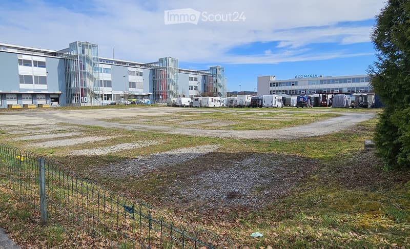 Modern multi-story building with glass facades and several parked vehicles, including caravans and trucks, in an open area with a fence in the foreground.