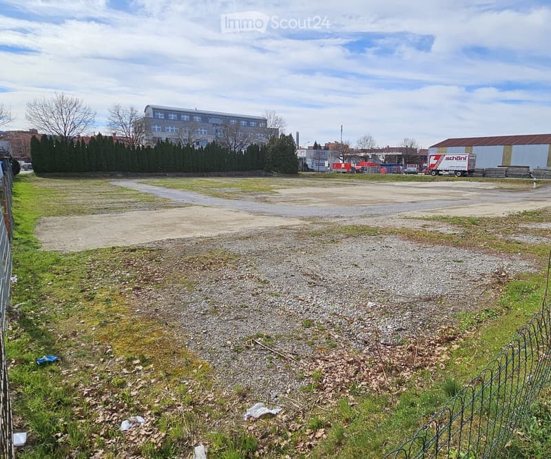 An open, mostly empty lot with patches of grass and gravel, surrounded by a fence and some trees. In the background, there are buildings and a few vehicles.