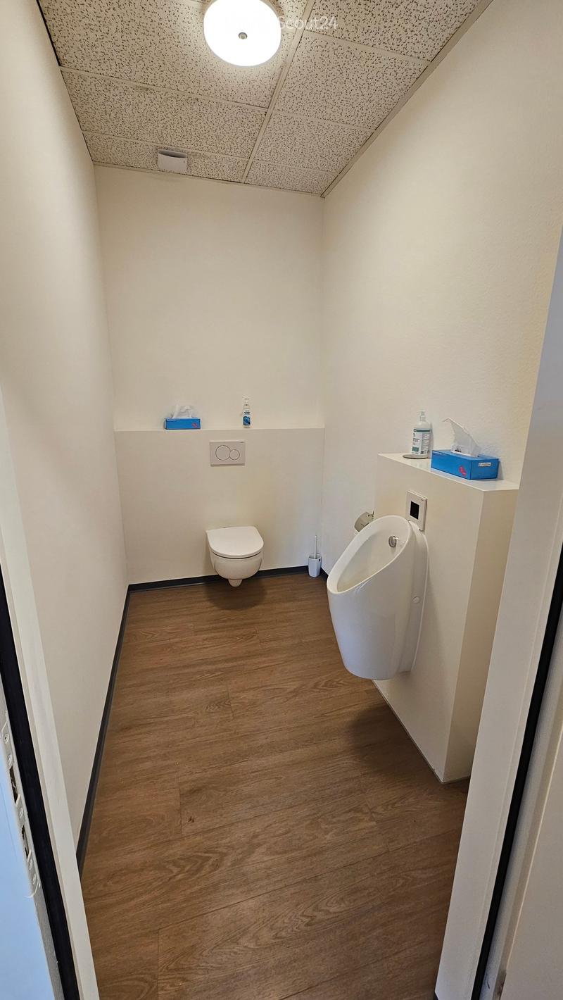Modern bathroom with a toilet, urinal, and wooden flooring. Includes a small shelf with a tissue box and bottle.