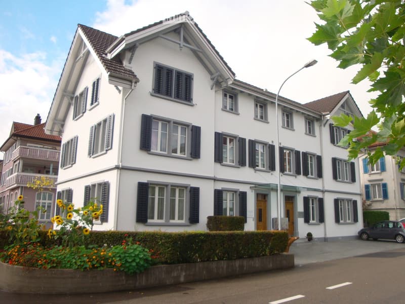 A multi-story white house with dark shutters, multiple windows, and a well-maintained garden in front.