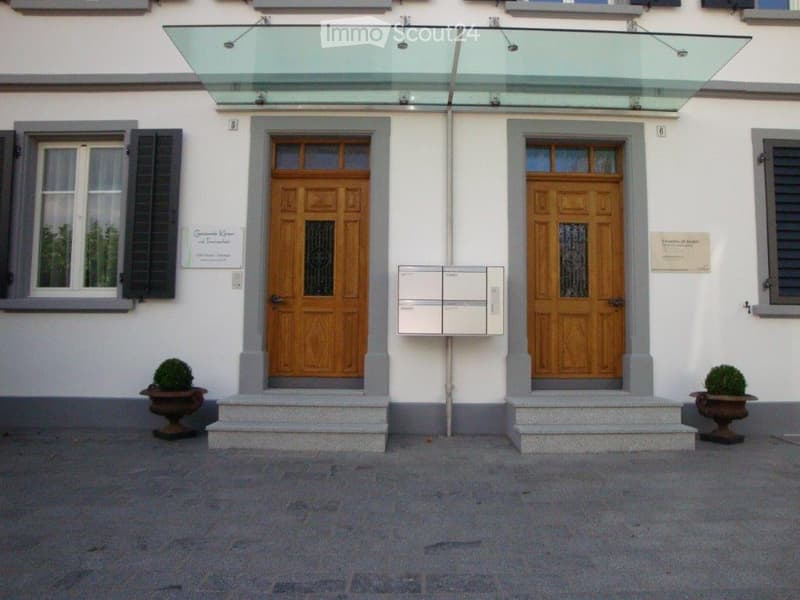 Entrance to a building with two wooden doors, a glass canopy, mailboxes, and potted plants on either side.