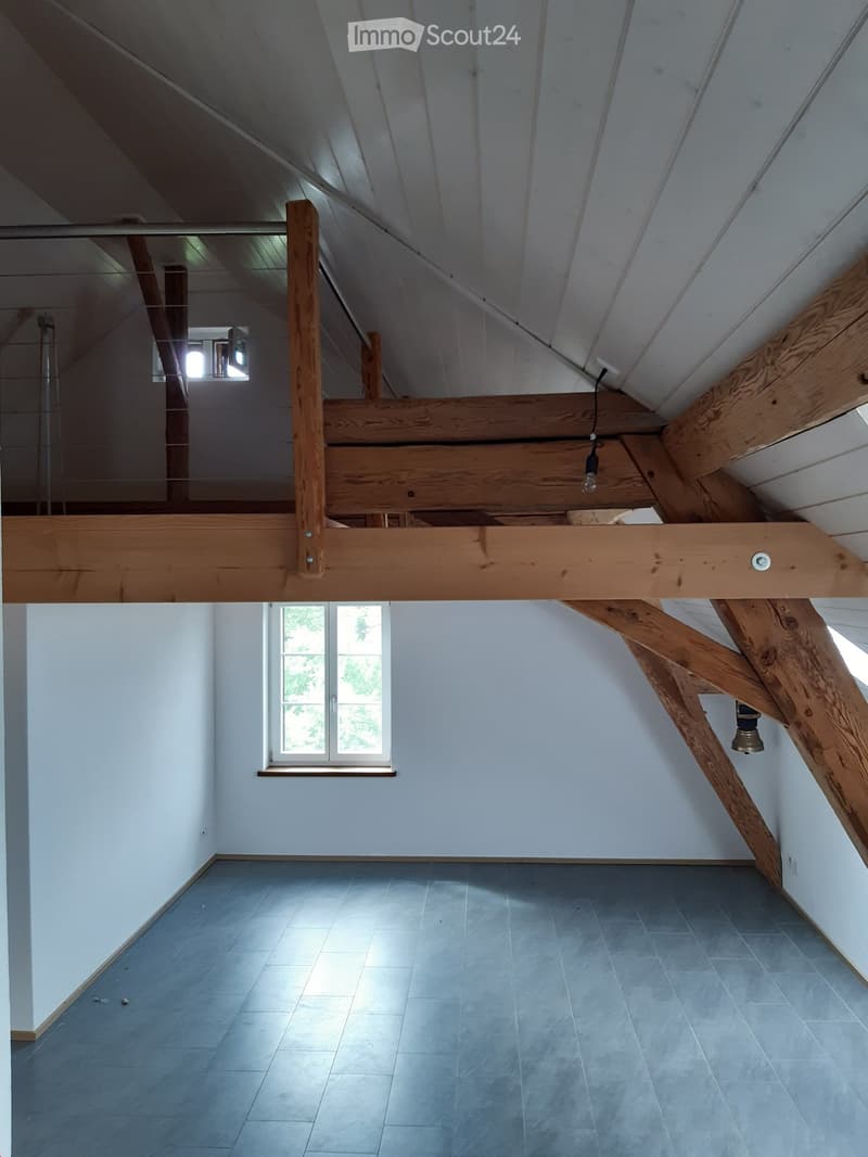 Empty room with grey tiled floor, white walls, and a large window letting in natural light. Exposed wooden beams and a glass railing are visible, indicating a loft or attic space.