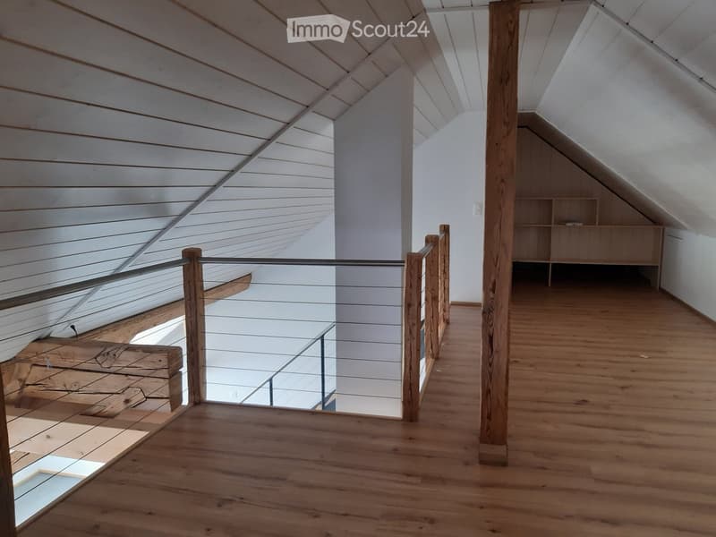 Spacious hallway with wooden flooring, white walls, and a glass railing leading to a lower level. The ceiling features exposed wooden beams and a slanted design, with built-in shelving on one side.