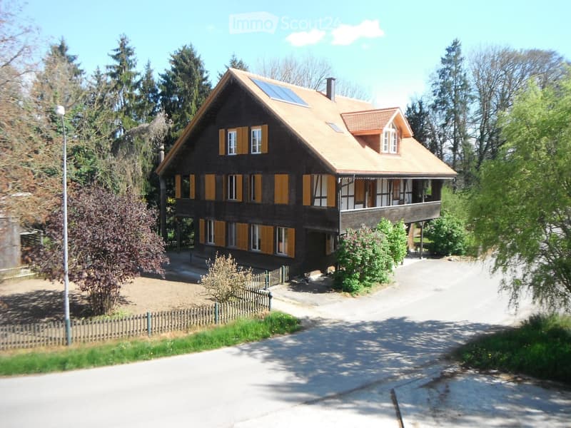 Two-story wooden house with a tiled roof, yellow shutters, and a small balcony on the upper level. The house is surrounded by trees and a wooden fence.