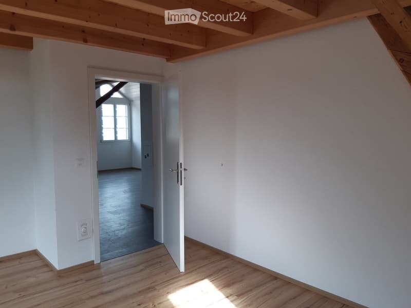 Empty room with wooden flooring, white walls, and exposed wooden beams on the ceiling. An open doorway leads to another room with a window and wooden floor.