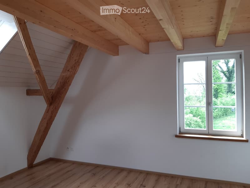 Empty attic room with wooden beams, exposed wooden ceiling, and a large window showing greenery outside.