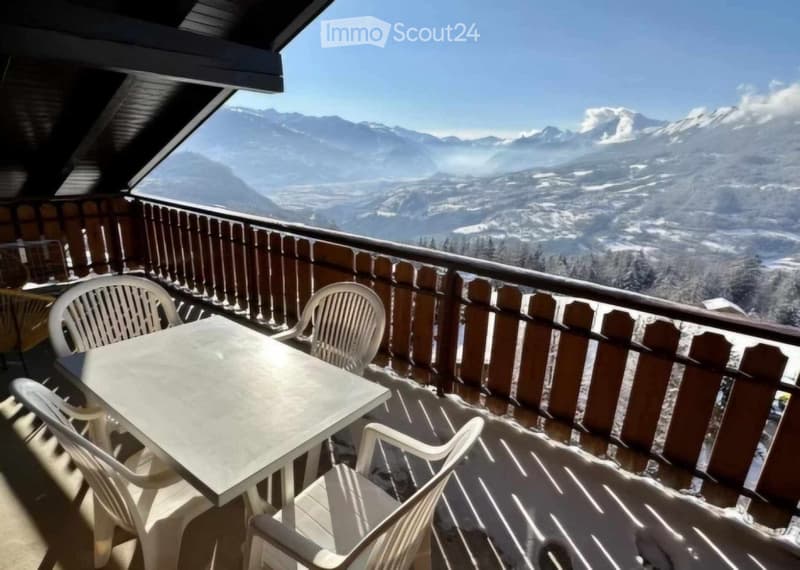 Balcony with wooden railing, overlooking a mountain range, equipped with a table and chairs.