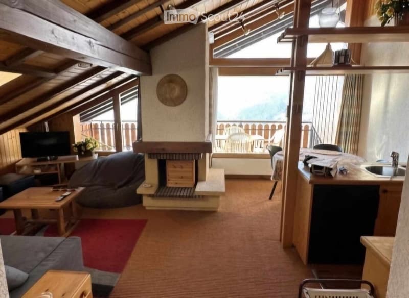 Cozy attic room with exposed wooden beams, fireplace, sofa, coffee table, television, and a small kitchen area with a sink. Large windows and a balcony offer a scenic view.