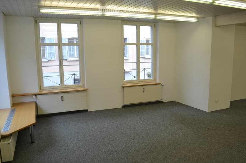 empty room, two large windows, wooden desk, white walls, carpeted floor, ceiling lights