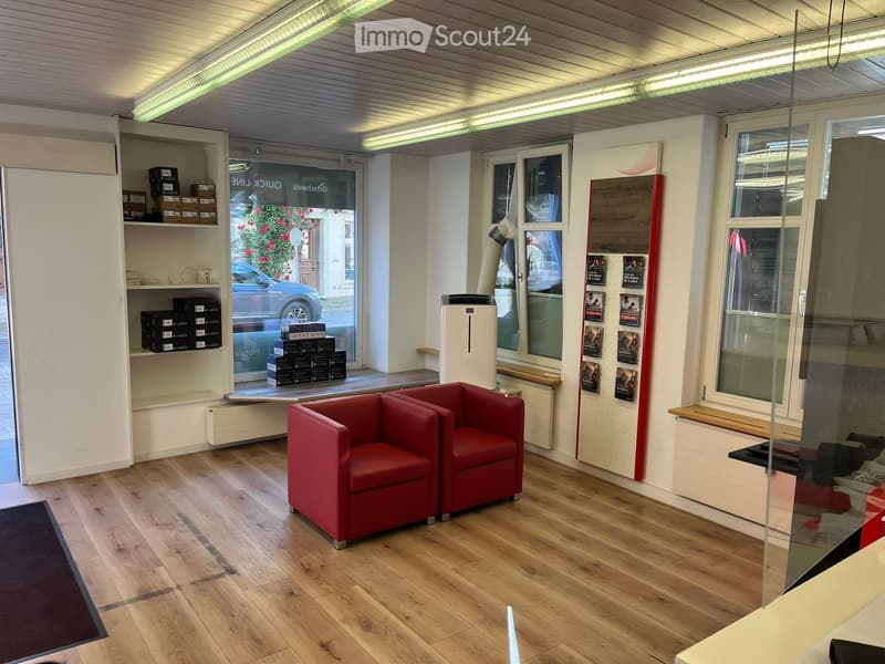 A modern, well-lit room with wooden flooring, two red armchairs, a shelf with boxes, an air conditioner, and posters on the wall. Large windows provide natural light and a view of the street.