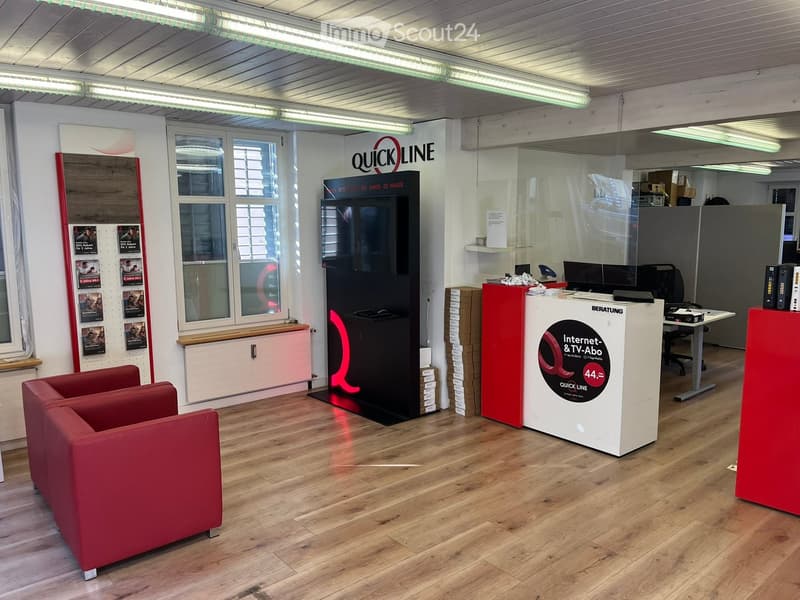 Modern office space with wooden flooring, red sofa, large screen, and a reception desk.