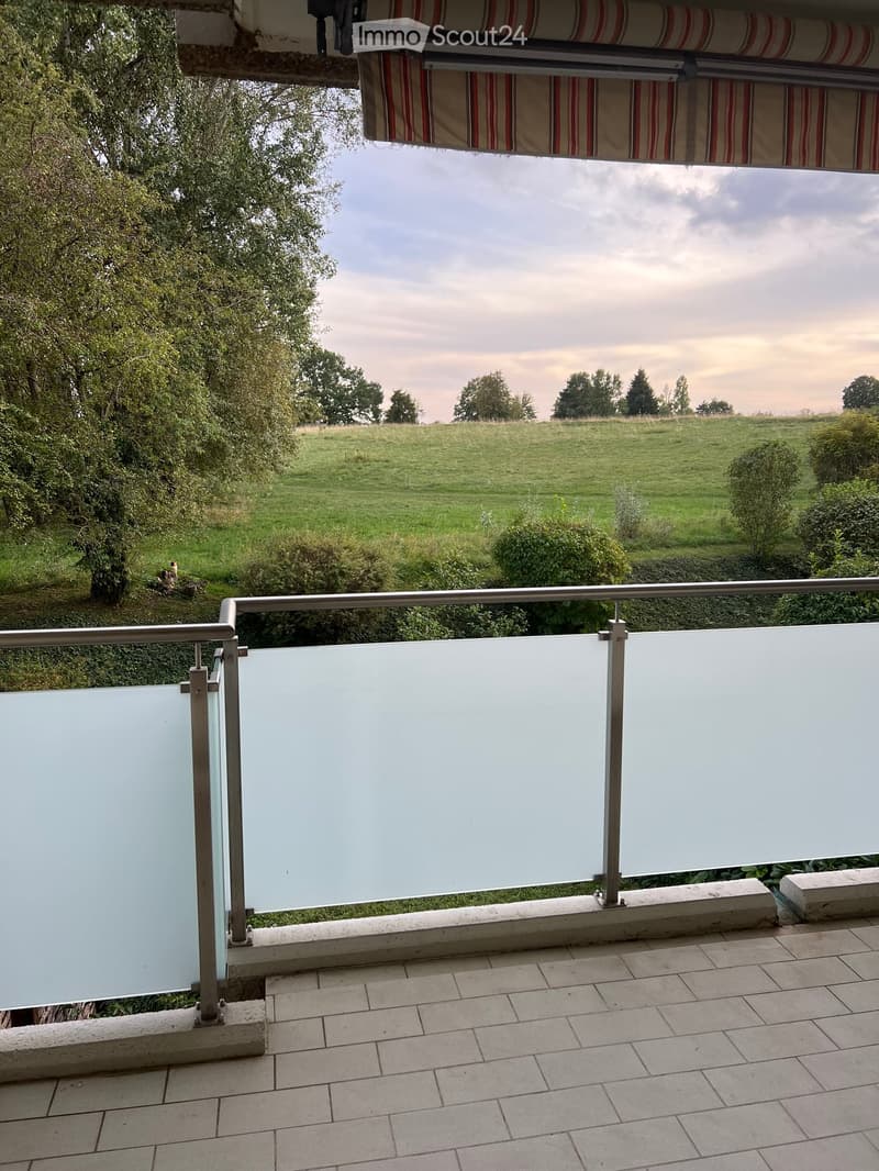 Balcony with glass panels, metal railing, tiled floor, and a view of a grassy field and trees.