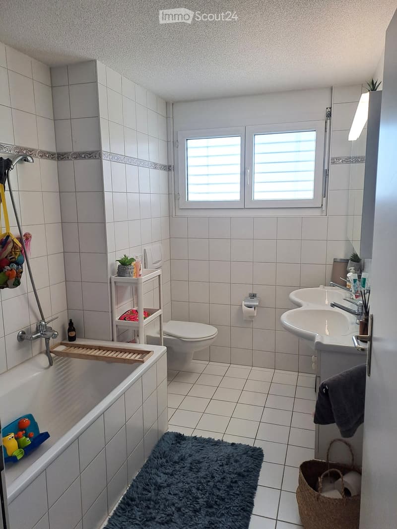 Bathroom with bathtub, toilet, double sink, shelves, rug, and a basket with towels.