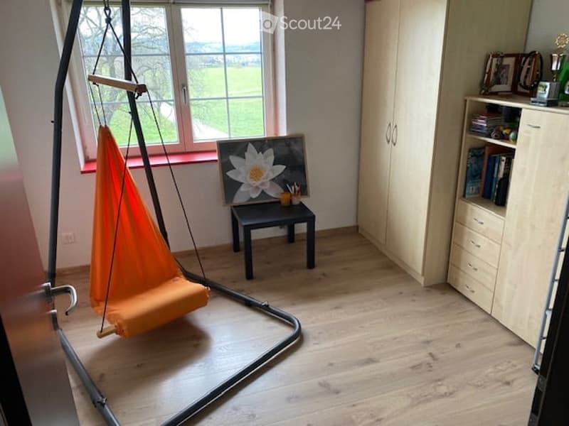 A cozy, well-lit room with wooden flooring, a hanging orange hammock, a small black stool with a flower painting, a tall white cabinet, and a bookshelf filled with books and decorative items.