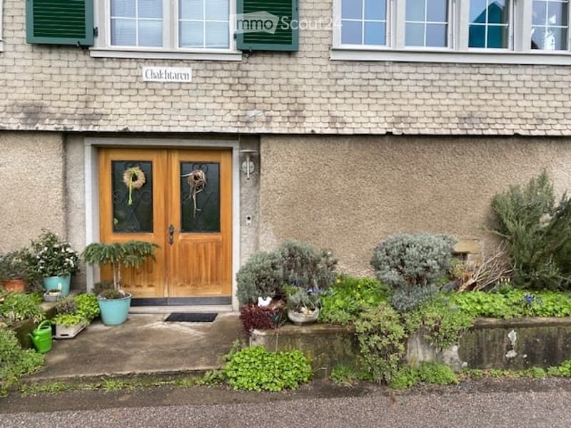 Front view of a two-story house with a wooden double door, green shutters, and a small garden with potted plants and shrubs.