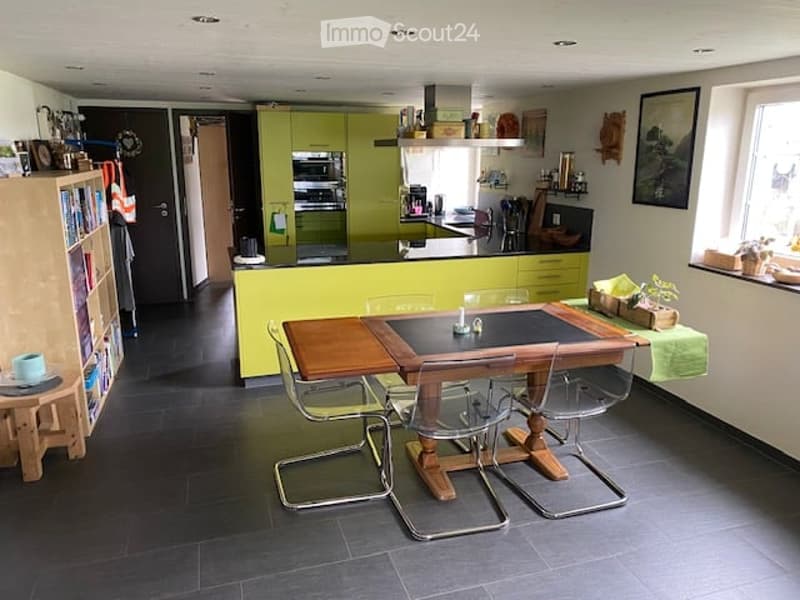 Bright kitchen and dining area with modern green cabinets, a wooden dining table with four chairs, and a bookshelf with books and decorative items.