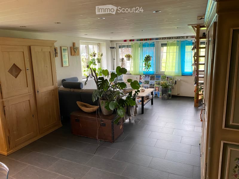 Bright living room with dark tiled floor, large windows, and wooden ceiling. Features a sofa, coffee table, potted plants, and a wooden cabinet.
