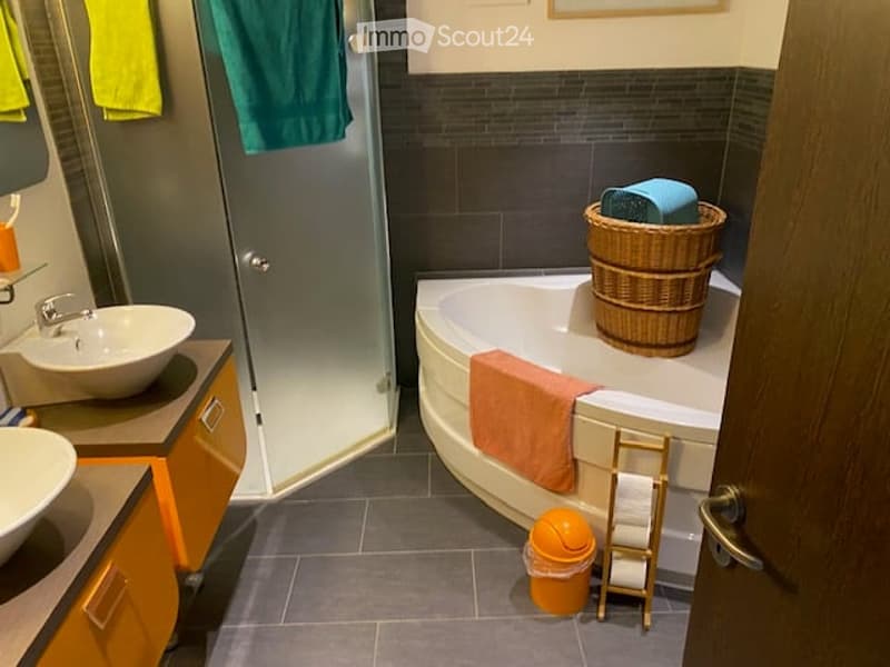 Modern bathroom with a white bathtub, two sinks, a glass shower enclosure, and a wicker basket on the edge of the tub. The space features dark grey tiles and warm wood accents.