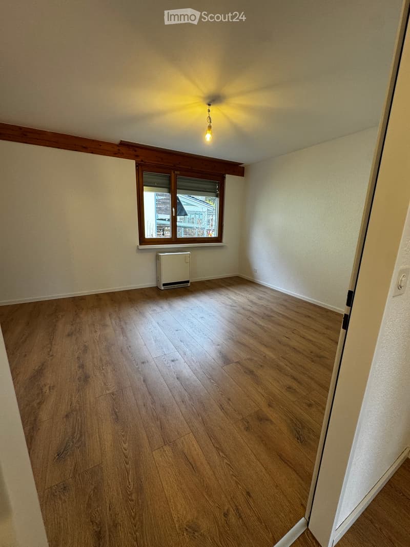 Empty room with wooden flooring, white walls, a single window with wooden frame, and a small white radiator under the window. The ceiling has a simple light fixture.