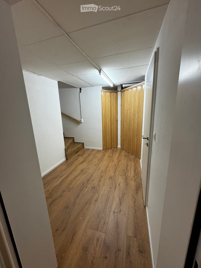 Empty hallway with wooden flooring, white walls, and a staircase leading upwards. Wooden doors are visible at the end of the hallway.