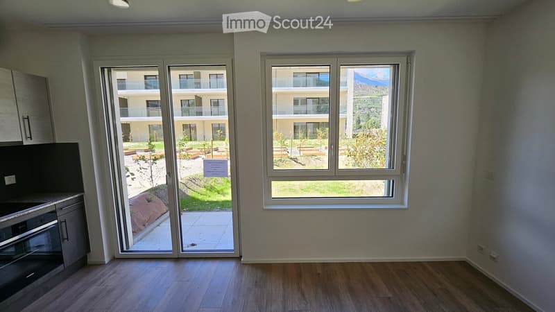 Empty room with wooden flooring, large windows, and sliding glass doors leading to a balcony area. The walls are painted in a light neutral color, and the room has modern cabinetry on one side.
