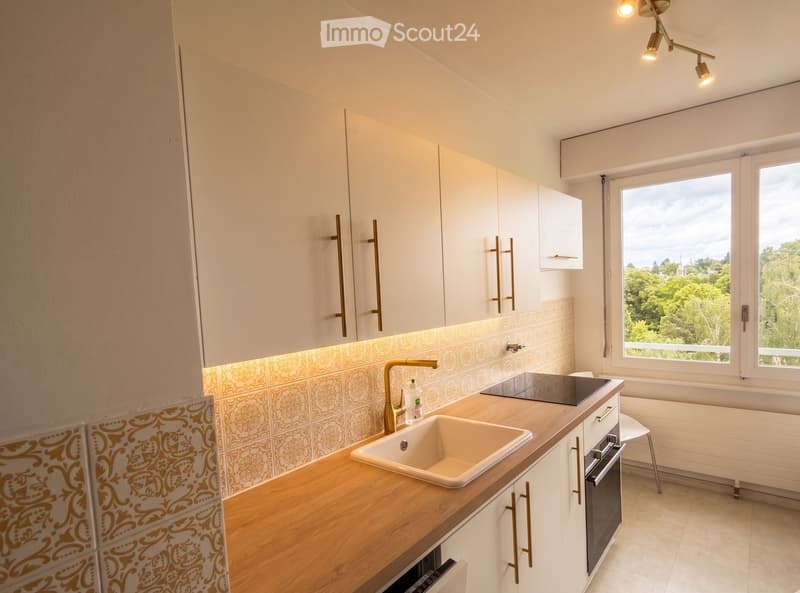 Modern kitchen with white cabinets, wooden countertop, sink, faucet, and built-in oven. Features decorative tiles and large windows with a view of greenery.