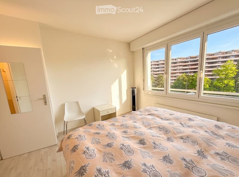 Bright bedroom with large windows offering a view of the city and greenery. Features a bed with a patterned duvet, a white chair, a nightstand, a mirror, and a radiator.