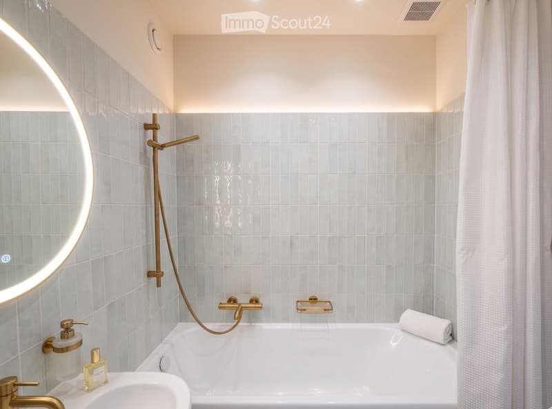 Modern bathroom with a bathtub, gold fixtures, a round mirror, and a towel on the edge of the tub.