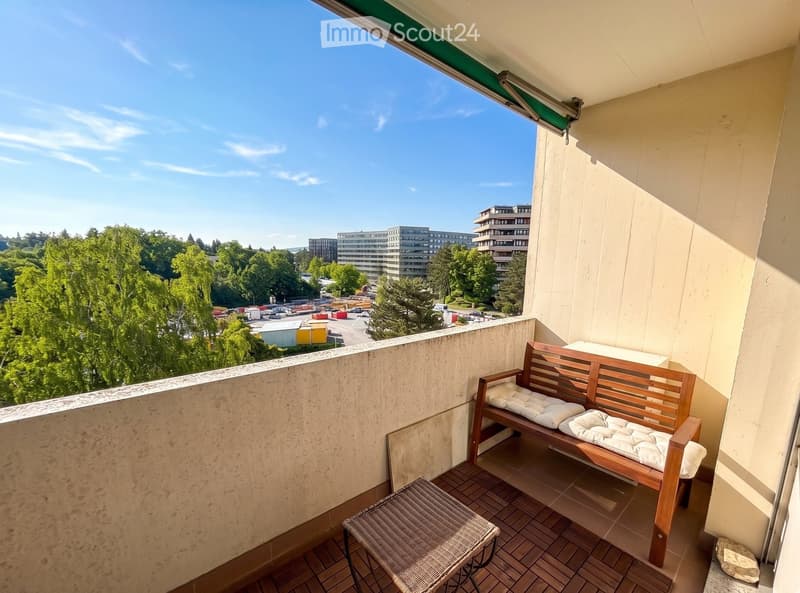 Balcony with wooden bench, cushioned seat, and wicker stool, overlooking a cityscape with trees and buildings.