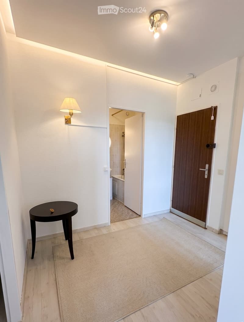 Bright hallway with wooden flooring, white walls, and recessed lighting. Features a small round table with a decorative item, a wall-mounted lamp, and an open doorway leading to a bathroom with a bathtub and shower area.