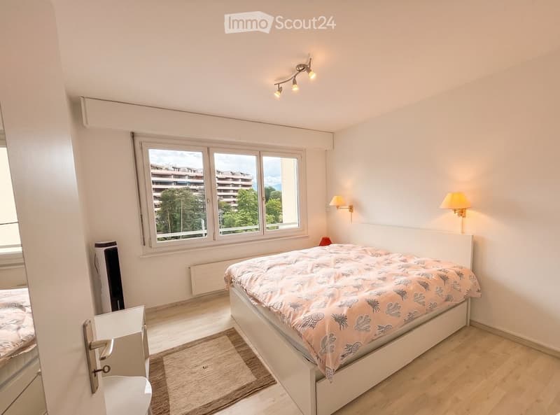 Modern bedroom with a large bed, white walls, wooden flooring, and large windows offering a view of buildings and greenery.