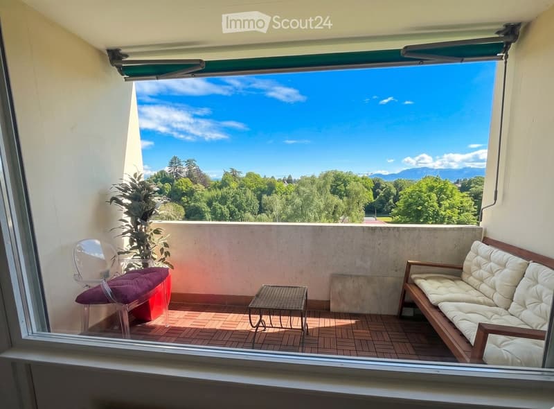 Balcony with wooden decking, white sofa, small table, chair, and potted plant, overlooking a green landscape with trees and mountains.