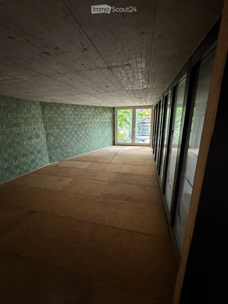 Modern, minimalist hallway with concrete ceiling and tiled walls, leading to an open doorway with glass panels.