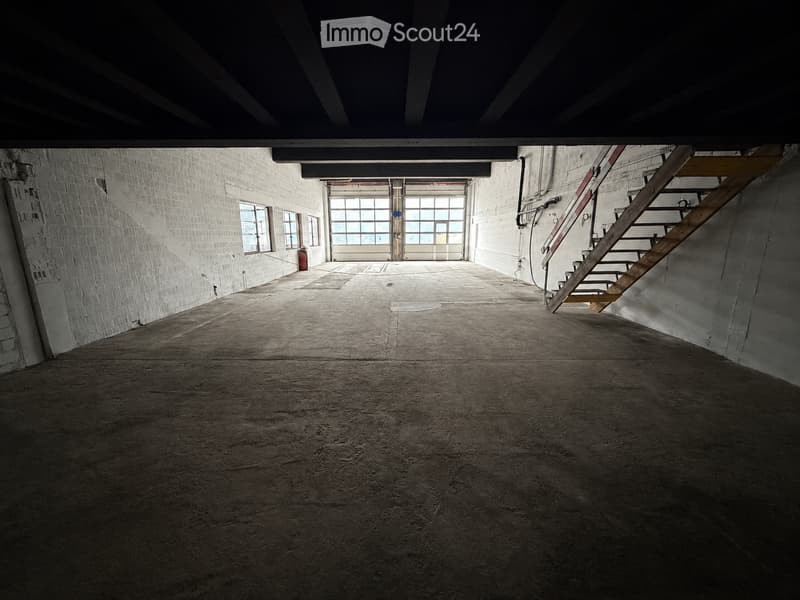 Empty industrial-style room with exposed brick walls, concrete floor, large windows, and a wooden staircase leading upwards.