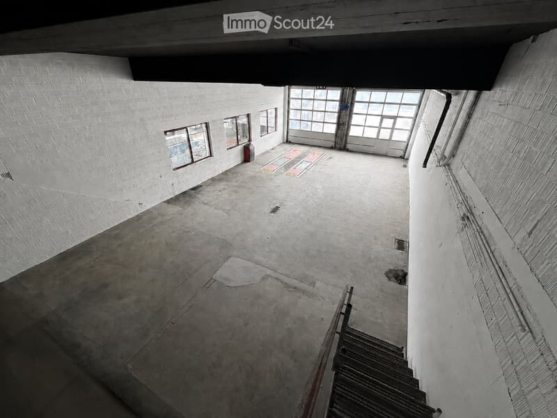 Empty room with white brick walls, concrete floor, large windows, and a metal staircase leading downwards.