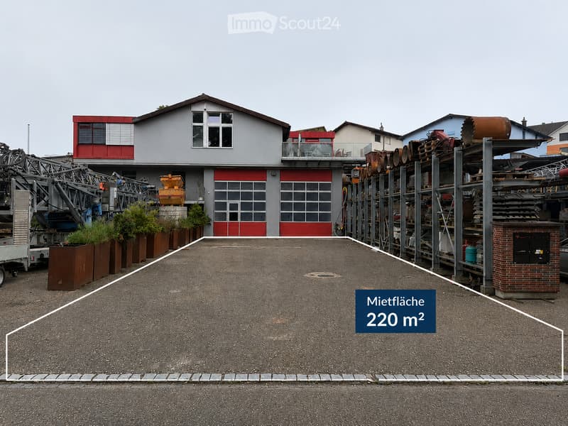 A modern industrial-style building with a grey facade, red accents, and large garage-style doors. The surrounding area includes storage racks, machinery, and potted plants.