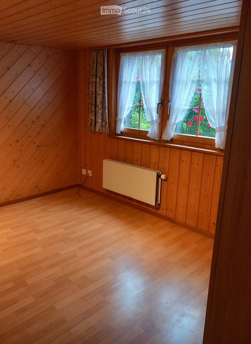 Empty room with wooden walls and floor, two windows with white curtains, radiator, and visible electrical outlets.