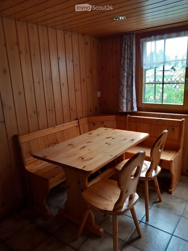 A cozy wooden room with a table, bench seating, and chairs, featuring wooden walls and a window with a view of greenery.