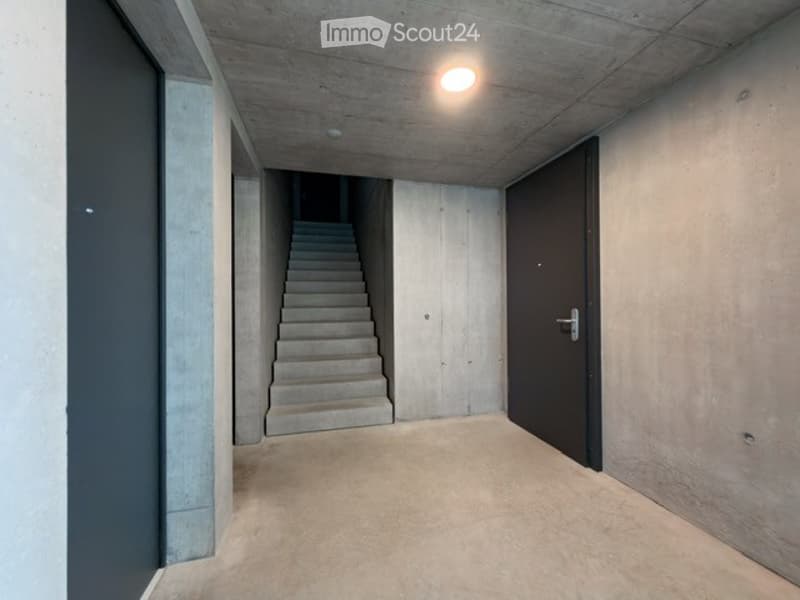 Minimalist concrete hallway with a staircase leading upwards, recessed lighting, and a closed door on the right.